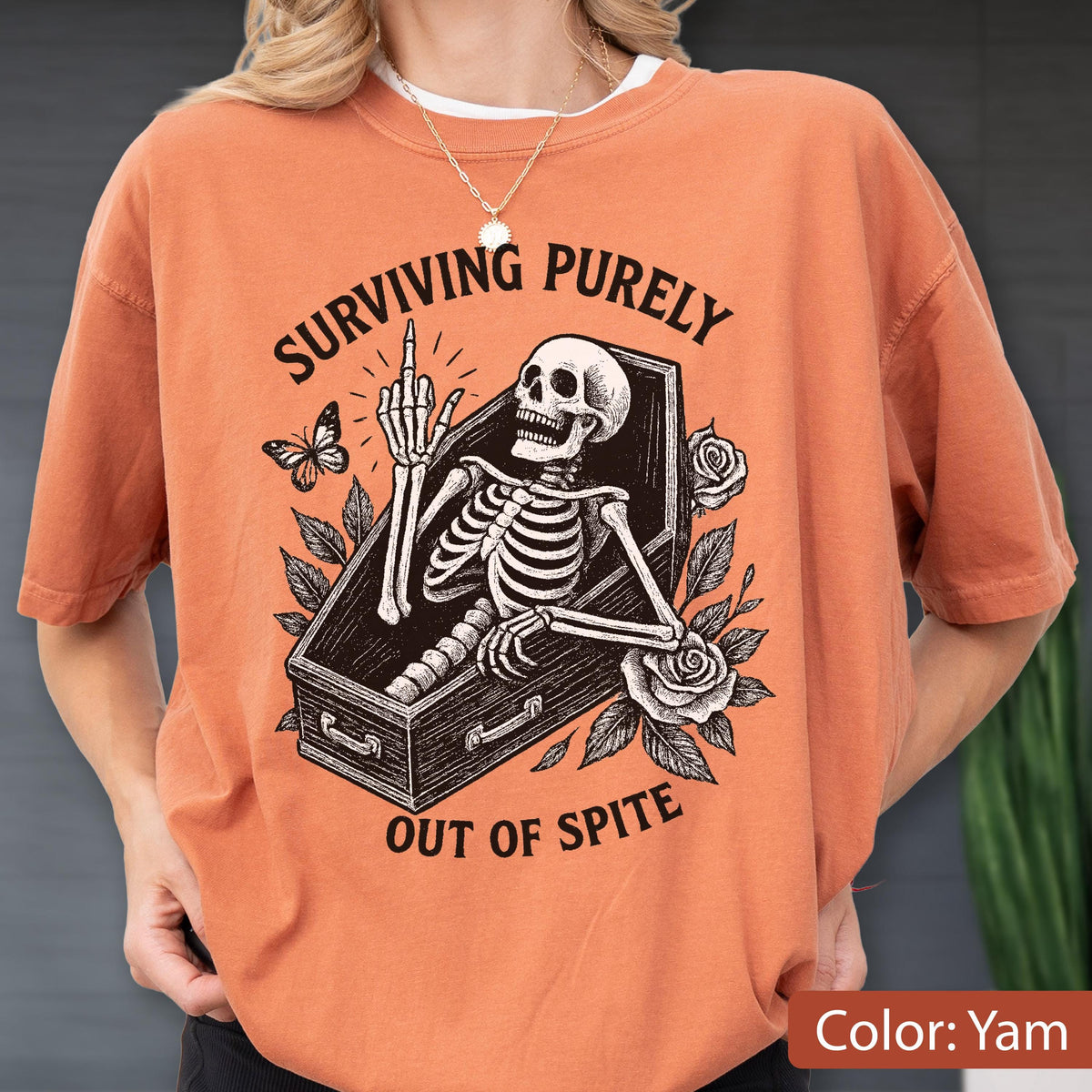 May include: A yam-colored t-shirt featuring a black graphic of a skeleton inside a coffin, giving the middle finger. The text reads "SURVIVING PURELY OUT OF SPITE" in a curved font. The design includes a butterfly and two roses with leaves.