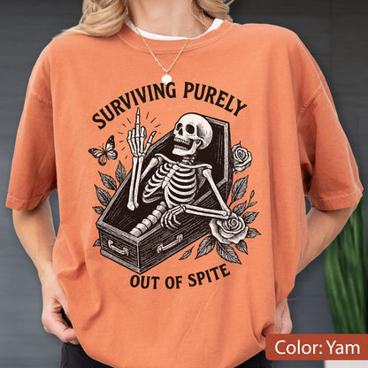 May include: A yam-colored t-shirt featuring a black graphic of a skeleton inside a coffin, giving the middle finger. The text reads "SURVIVING PURELY OUT OF SPITE" in a curved font. The design includes a butterfly and two roses with leaves.