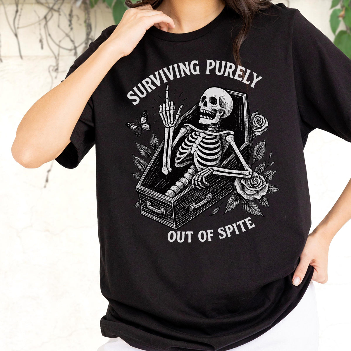 May include: A black t-shirt featuring a white graphic of a skeleton inside a coffin, giving the middle finger. The text reads "SURVIVING PURELY" above and "OUT OF SPITE" below, surrounded by illustrations of a butterfly and two roses.