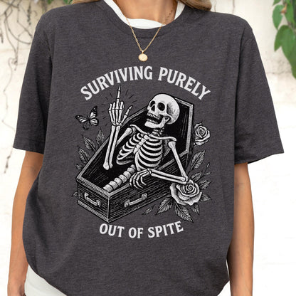 May include: A dark charcoal gray t-shirt featuring a white graphic print of a skeleton lying in a coffin, giving the middle finger. The design includes roses, a butterfly, and the text "SURVIVING PURELY OUT OF SPITE" in a bold, arched font.