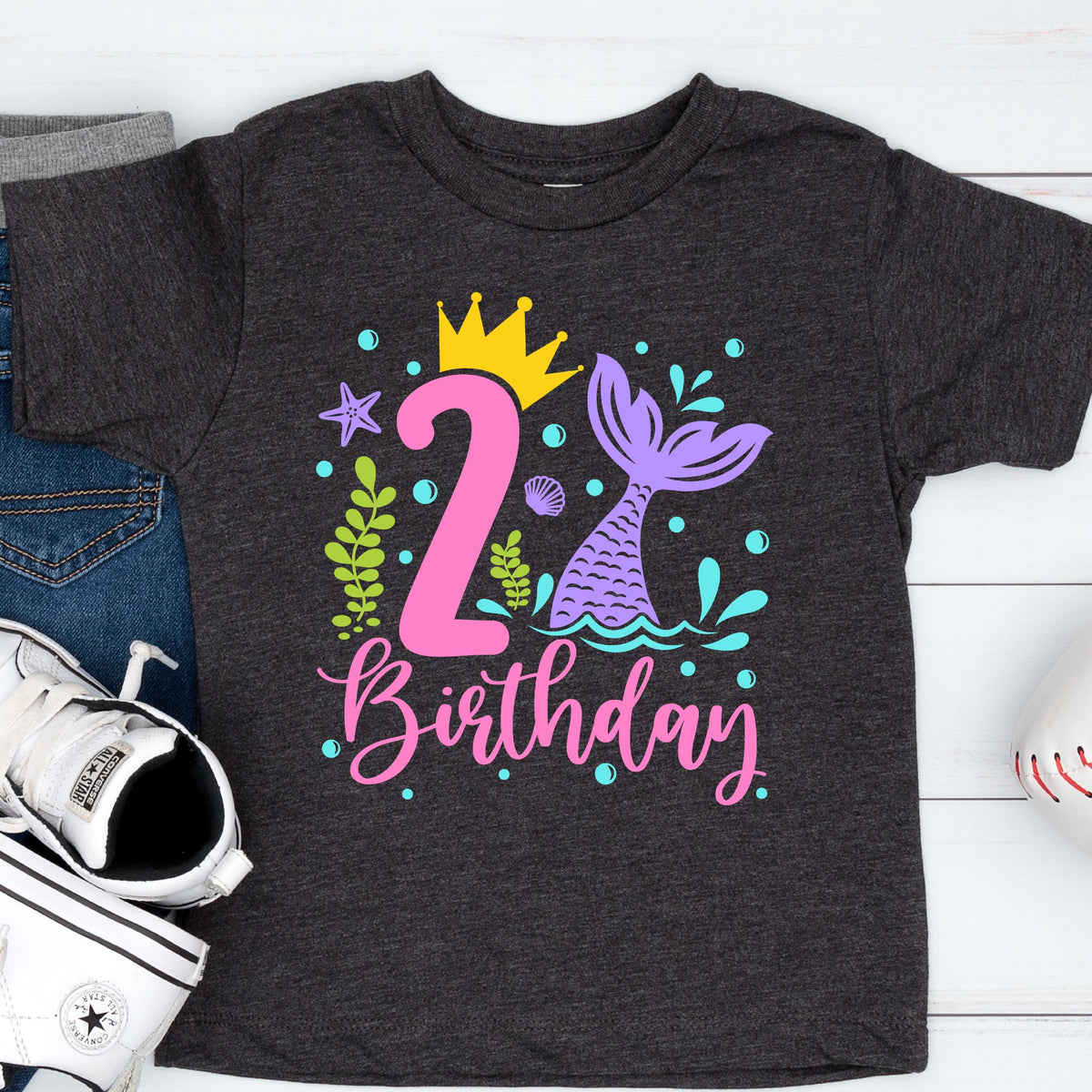 May include: Dark gray toddler t-shirt with a colorful mermaid-themed birthday design. The design includes a pink number 2, a yellow crown, a purple mermaid tail, and the word "Birthday" in pink script.