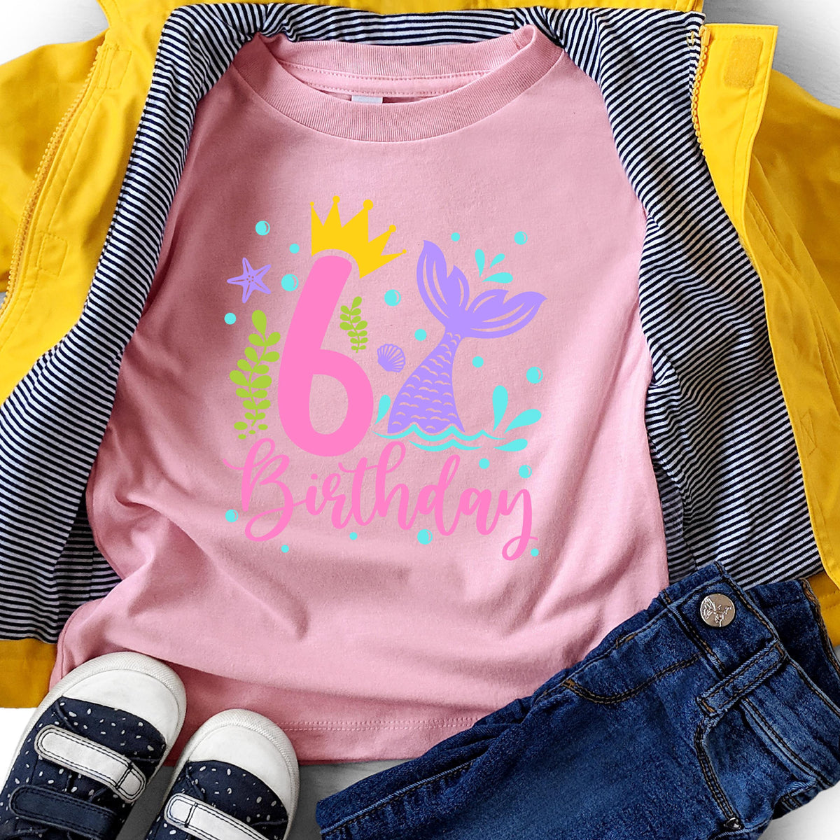 May include: A pink t-shirt with a mermaid theme, featuring a large number "6" and the word "Birthday" in a script font. The design includes a mermaid tail, crown, and aquatic elements. The shirt is displayed with a yellow jacket and blue jeans.