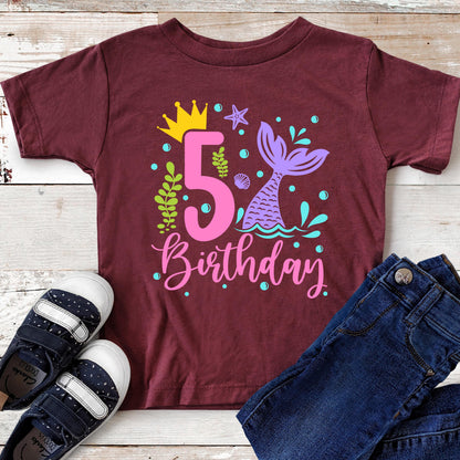 May include: Burgundy-colored children's t-shirt with a colorful mermaid-themed design. The graphic includes a large pink number 5, a mermaid tail, and the word "Birthday". A pair of navy blue shoes and blue jeans are next to the shirt.