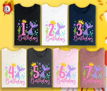 May include: A collection of seven t-shirts in various colors, each featuring a mermaid tail design with a number and the word "Birthday." The shirts are black, white, navy, cream, charcoal, pink, and mauve. The designs include a gold crown and aquatic elements.