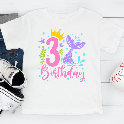 May include: White t-shirt with a colorful mermaid-themed design for a third birthday. The design includes a pink number 3, a mermaid tail, a crown, and the word "Birthday" in pink script.
