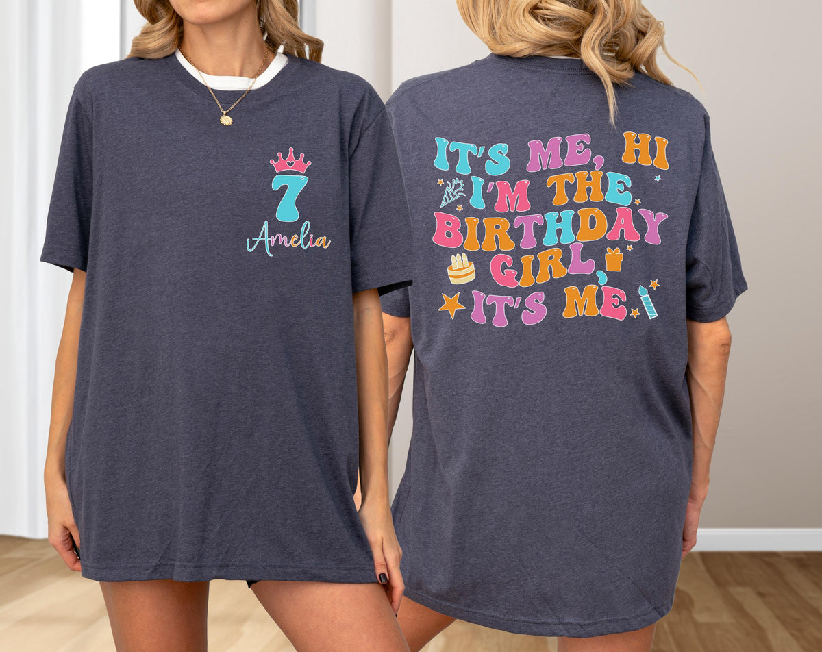 May include: A dark heather gray t-shirt featuring a front graphic with a pink crown, the number 7, and the name Amelia. The back displays colorful retro-style text reading IT'S ME, HI I'M THE BIRTHDAY GIRL IT'S ME, with icons of a cake and candle.