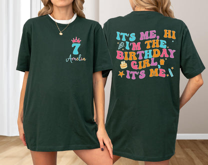 May include: A dark green t-shirt featuring a front graphic with a pink crown, the number 7, and the name Amelia. The back displays colorful retro-style text reading IT'S ME, HI I'M THE BIRTHDAY GIRL IT'S ME, accompanied by icons of a cake, gift, and candle.