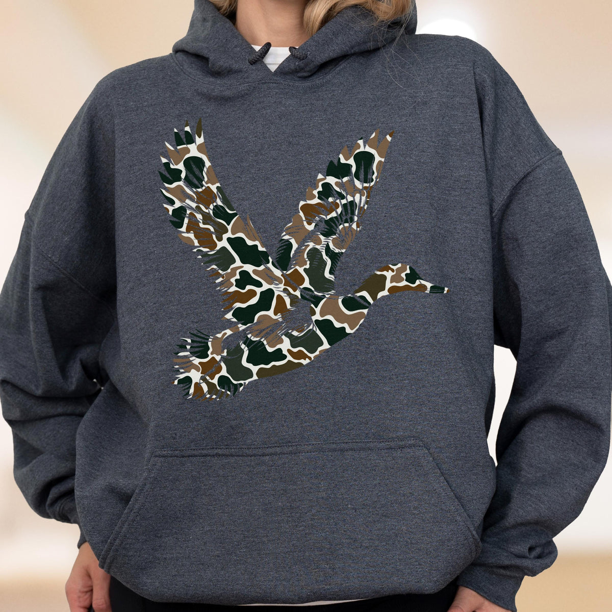 May include: A dark charcoal gray hooded sweatshirt featuring a large graphic of a flying duck on the chest. The duck silhouette is filled with a camouflage pattern consisting of brown, dark green, black, and white organic shapes and patches.