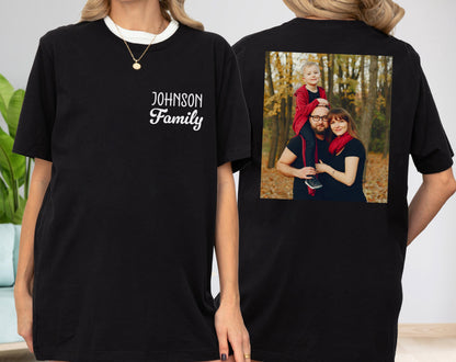 May include: Black t-shirt with a white collar and the text "Johnson Family" on the front. The back of the shirt features a family photo. The family is wearing black shirts and posing in front of a background of trees.