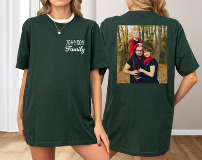 May include: Dark green t-shirt with the text "JOHNSON Family" in white script on the front. The back of the shirt features a family photo in a square frame. The family photo shows a man, woman, and child in a forest setting.