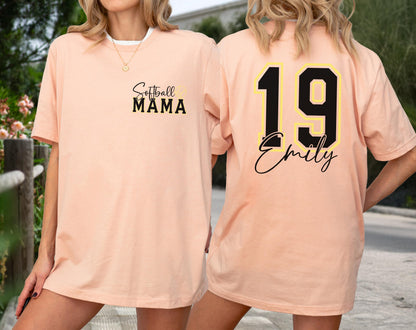 May include: Peach-colored t-shirt with the text "Softball MAMA" on the front. The back of the shirt features the number "19" in black with a yellow outline, and the name "Emily" in a script font.