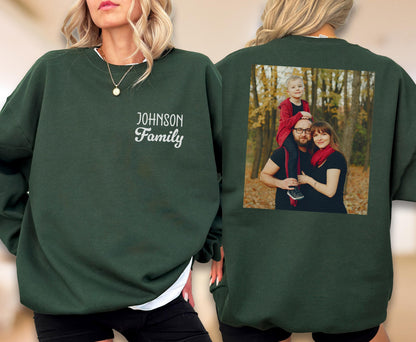 May include: A dark green sweatshirt with the text "JOHNSON Family" in white script on the front. The back of the sweatshirt features a family photo in a rectangular frame. The family photo shows a man, woman, and child.