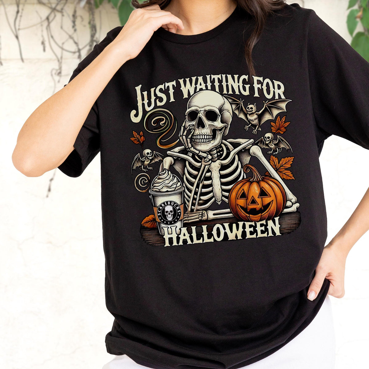 May include: Black t-shirt with a Halloween-themed graphic. The design features a skeleton with a coffee cup and a jack-o'-lantern, with the text "Just Waiting For Halloween". Bats and fall leaves complete the design.