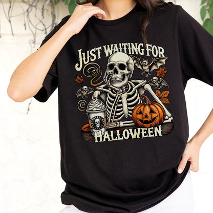 May include: Black t-shirt with a Halloween-themed graphic. The design features a skeleton with a coffee cup and a jack-o'-lantern, with the text "Just Waiting For Halloween". Bats and fall leaves complete the design.