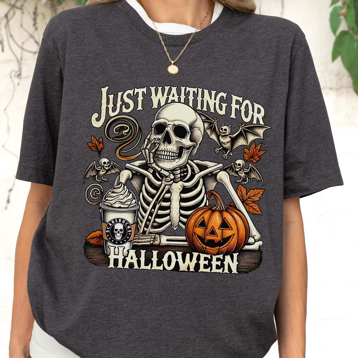 May include: Dark gray t-shirt with a Halloween-themed graphic. The design features a skeleton with a coffee cup, pumpkin, and bats, with the text "Just Waiting For Halloween". The shirt has a crew neck and short sleeves.