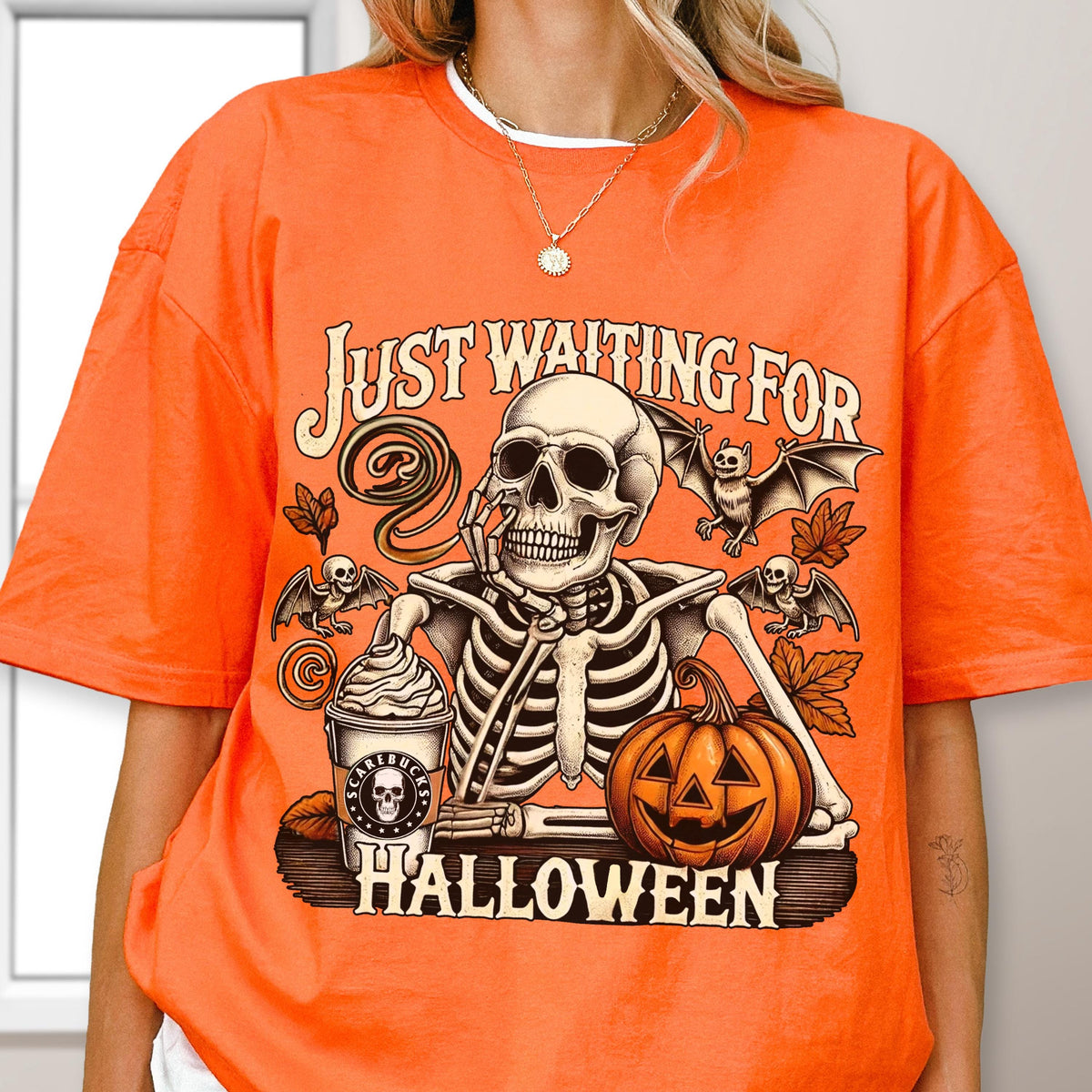 May include: Orange t-shirt with a Halloween-themed graphic. The design features a skeleton, bats, a pumpkin, and the text "JUST WAITING FOR HALLOWEEN". The skeleton is holding its chin, and a coffee cup is next to it.