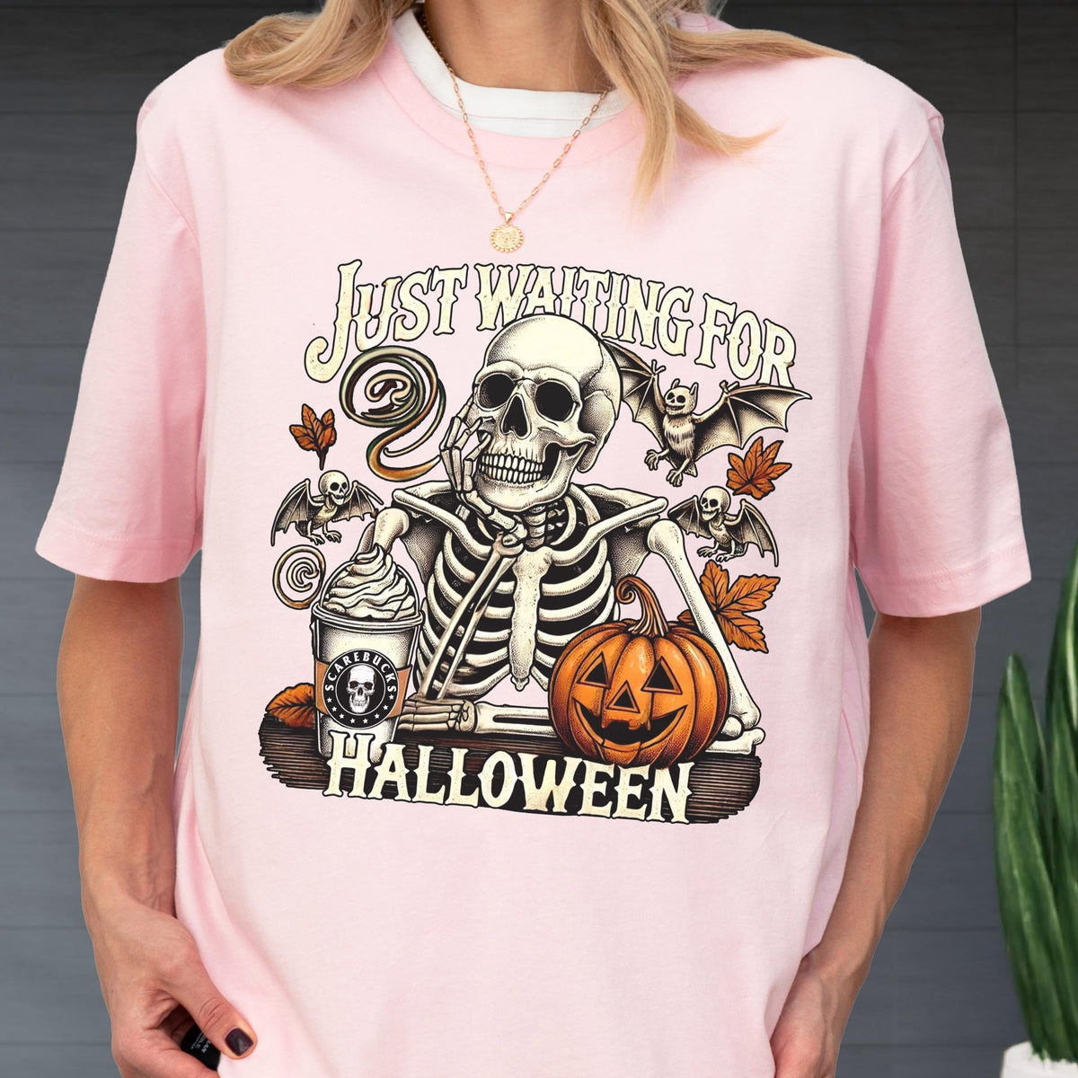 May include: Pink t-shirt featuring a skeleton with a coffee cup and pumpkin, with the text "Just Waiting For Halloween". The design includes bats and fall leaves. A fun, spooky design for the Halloween season.