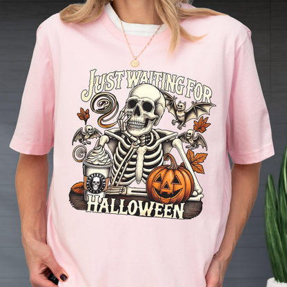 May include: Pink t-shirt featuring a skeleton with a coffee cup and pumpkin, with the text "Just Waiting For Halloween". The design includes bats and fall leaves. A fun, spooky design for the Halloween season.
