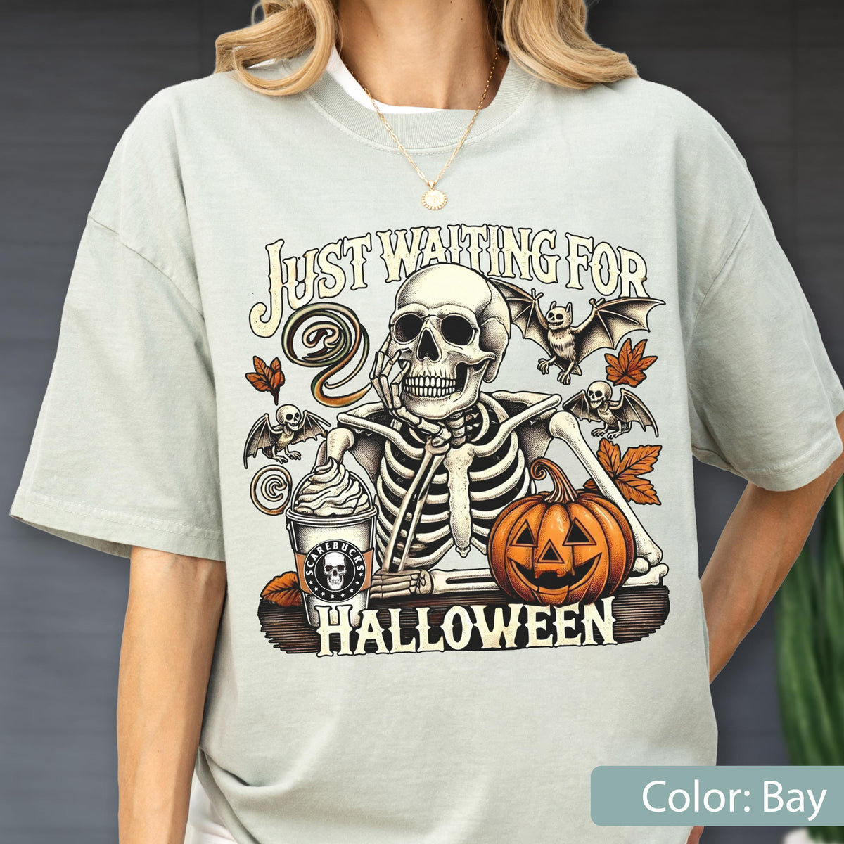 May include: A light sage green t-shirt featuring a graphic of a skeleton resting its chin on its hand next to a pumpkin and a coffee cup labeled Scarebucks. The text reads Just Waiting For Halloween, surrounded by bats and autumn leaves.
