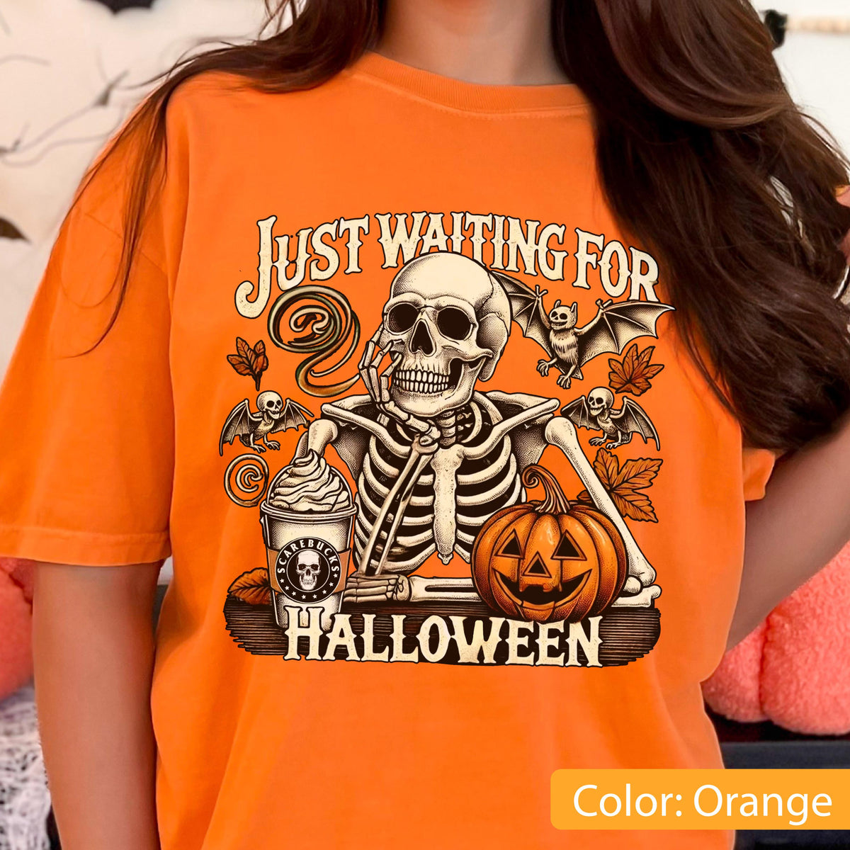 May include: An orange t-shirt featuring a graphic print of a skeleton resting its chin on its hand next to a pumpkin and a cup with a skull logo. The text reads "JUST WAITING FOR HALLOWEEN" with illustrations of bats and autumn leaves surrounding the skeleton.