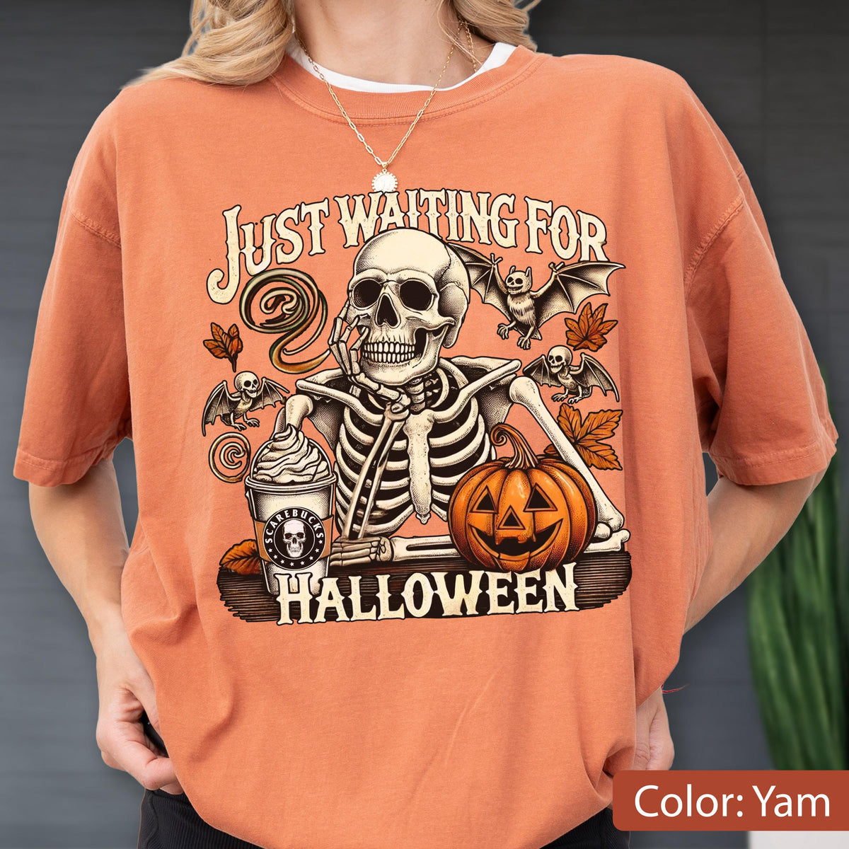 May include: A yam-colored t-shirt featuring a graphic of a skeleton resting its chin on its hand next to a pumpkin and a coffee cup labeled Scarebucks. The text reads Just Waiting For Halloween, surrounded by bats and autumn leaves.