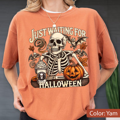 May include: A yam-colored t-shirt featuring a graphic of a skeleton resting its chin on its hand next to a pumpkin and a coffee cup labeled Scarebucks. The text reads Just Waiting For Halloween, surrounded by bats and autumn leaves.