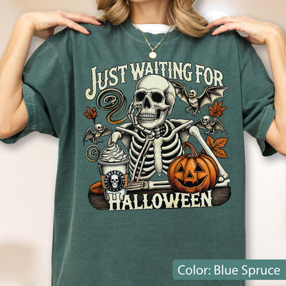 May include: A blue spruce t-shirt featuring a graphic of a skeleton resting its chin on its hand next to a pumpkin and a coffee cup. The text reads "JUST WAITING FOR HALLOWEEN". The design includes two bats and orange autumn leaves on a dark background.