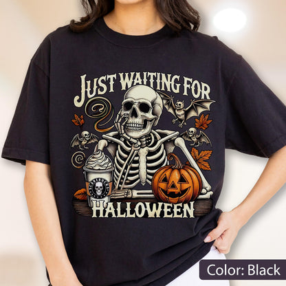 May include: A black t-shirt featuring a graphic of a skeleton resting its chin on its hand next to a pumpkin and a coffee cup labeled Scarebucks. The text reads Just Waiting For Halloween, surrounded by bats and autumn leaves in orange and brown.