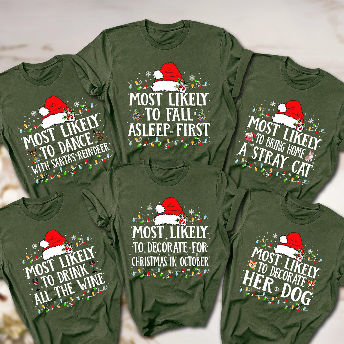May include: Six olive green t-shirts with various Christmas-themed phrases in white text, each topped with a Santa hat and string lights. Phrases include "Most Likely to Dance with Santa's Reindeer" and "Most Likely to Decorate Her Dog."