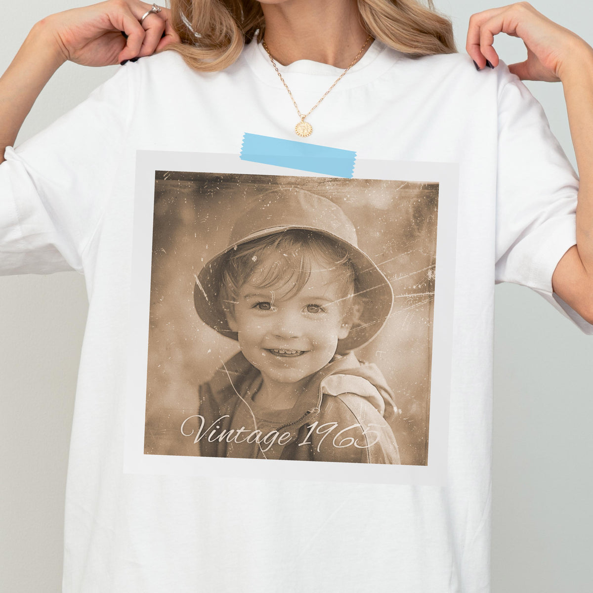 May include: White t-shirt with a sepia-toned photograph of a child wearing a hat. The photo is attached with blue tape and has the text "Vintage 1965" in cursive. The child is smiling.