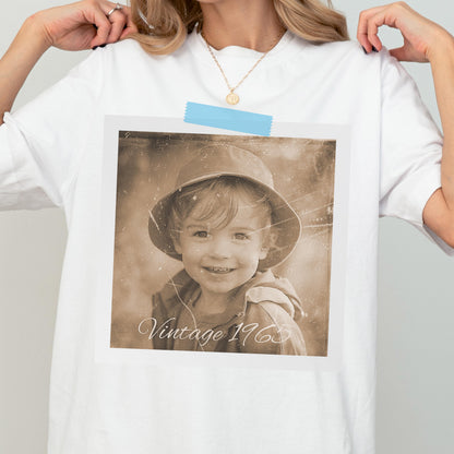 May include: White t-shirt with a sepia-toned photograph of a child wearing a hat. The photo is attached with blue tape and has the text "Vintage 1965" in cursive. The child is smiling.