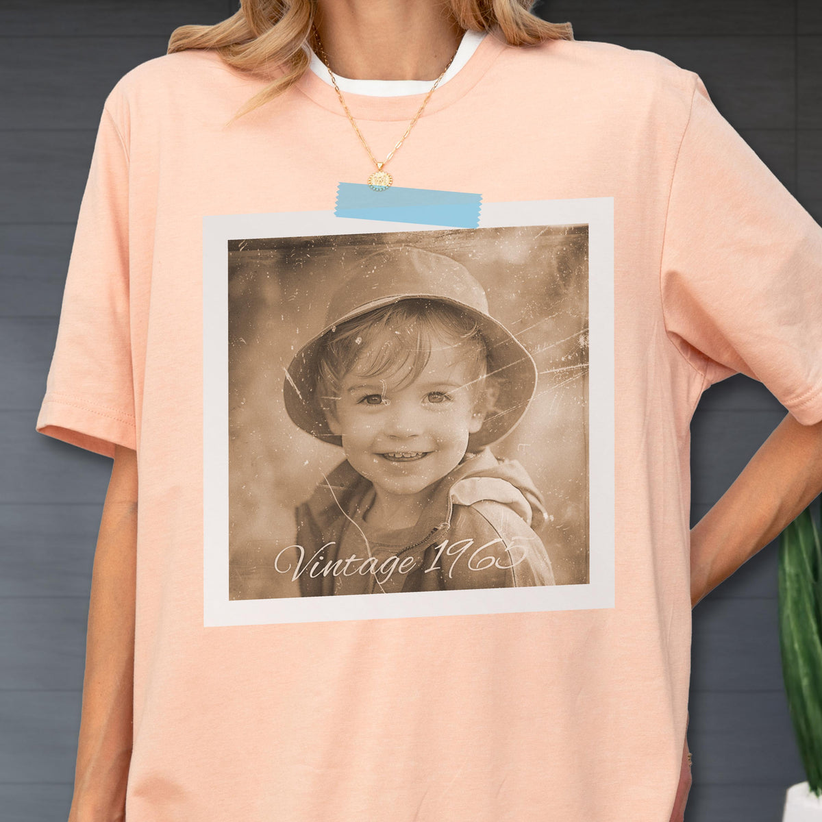 May include: Peach-colored t-shirt with a sepia-toned photo print of a child wearing a hat. The photo is attached with blue tape and has the text "Vintage 1963". The shirt has a white crew neck.