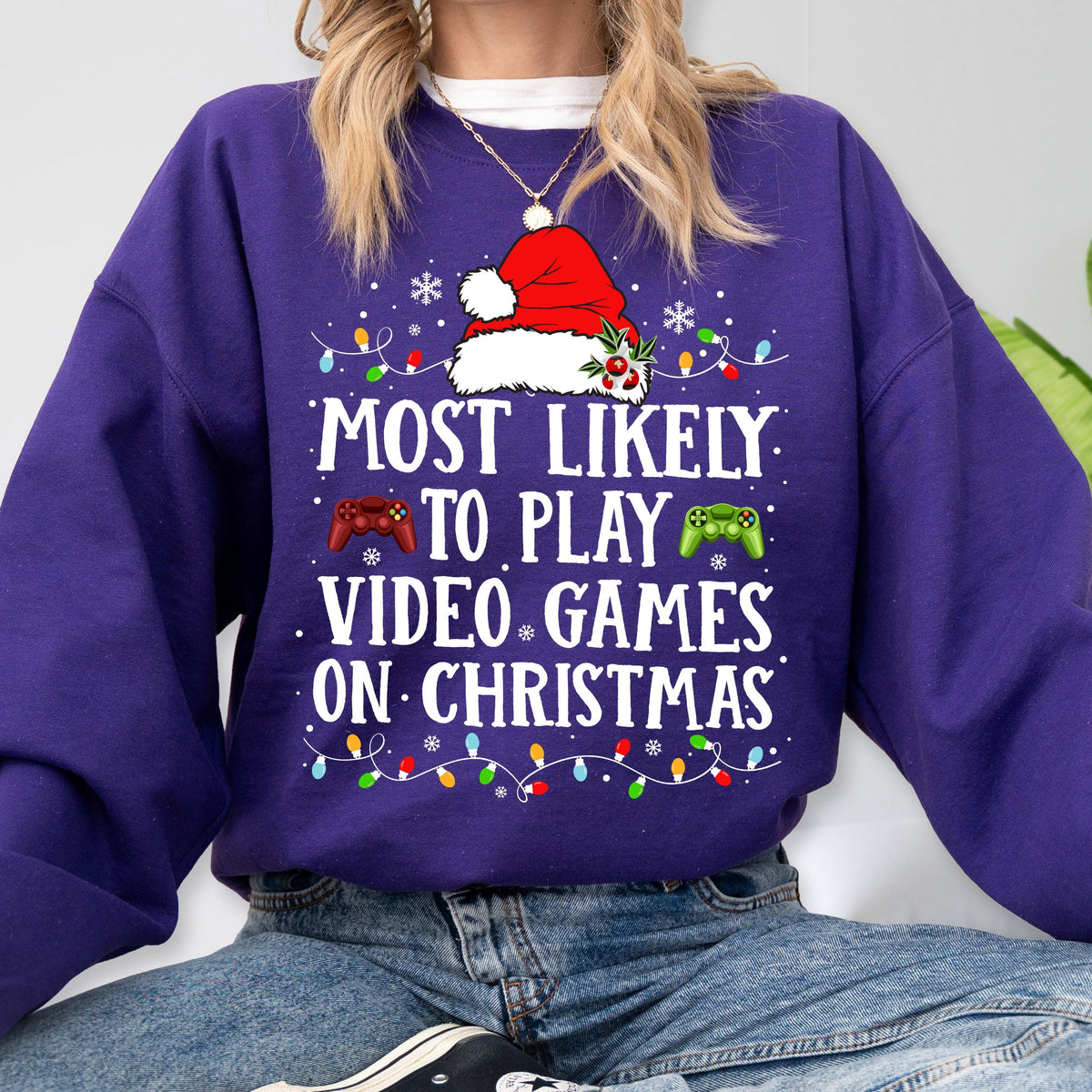 May include: A purple long-sleeve sweatshirt featuring a graphic of a Santa hat, red and green game controllers, and colorful Christmas lights. The text reads: MOST LIKELY TO PLAY VIDEO GAMES ON CHRISTMAS. The design includes small white snowflakes.