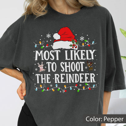 May include: A charcoal gray t-shirt featuring a graphic print of a red Santa hat above the white text "MOST LIKELY TO SHOOT THE REINDEER". The design includes small illustrations of reindeer, white snowflakes, and a string of colorful Christmas lights.