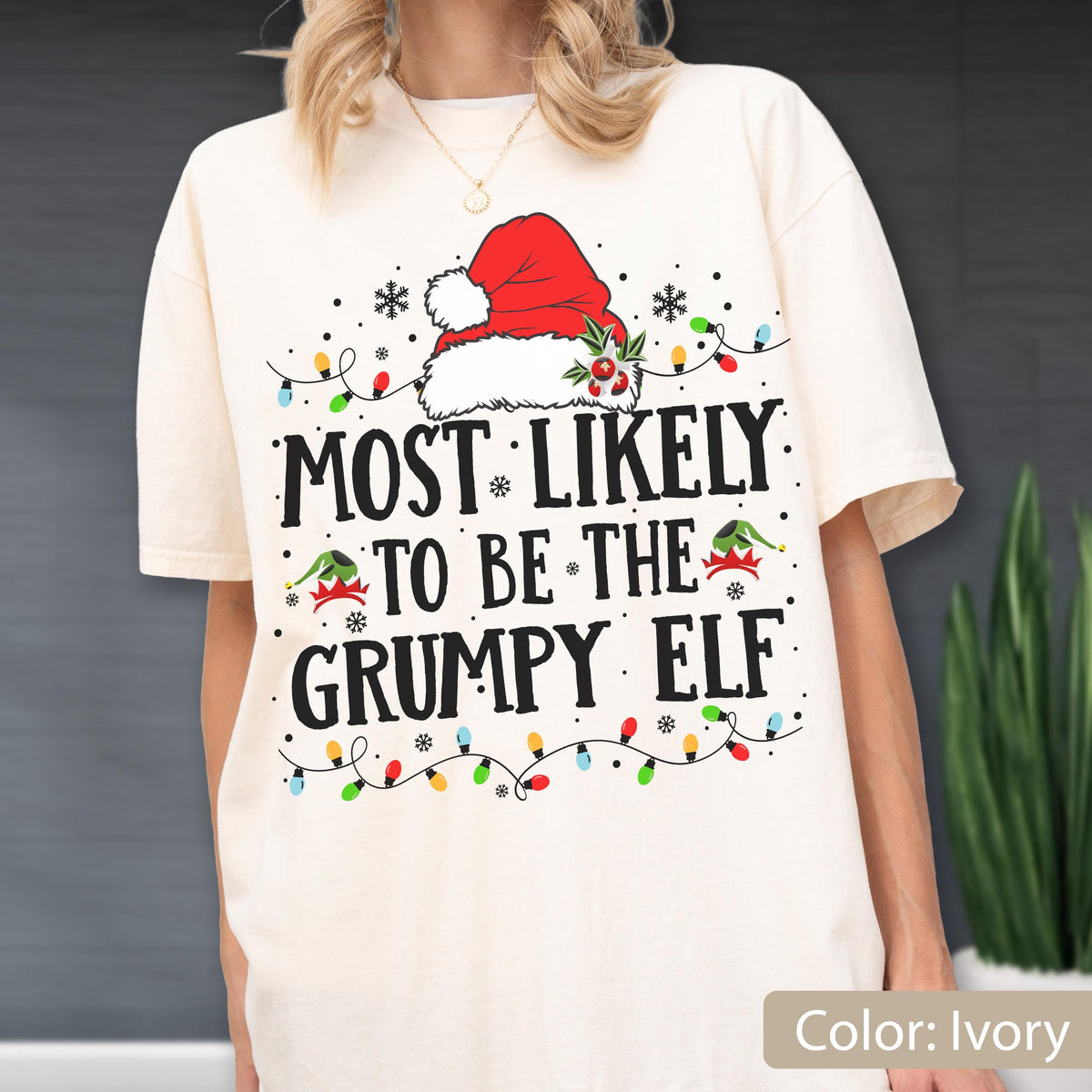 May include: Ivory-colored t-shirt with the text "MOST LIKELY TO BE THE GRUMPY ELF" in black. The design includes a red Santa hat, snowflakes, and colorful Christmas lights. The shirt is a festive holiday item.