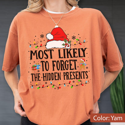 May include: A yam-colored t-shirt featuring a graphic of a Santa hat, a small teddy bear holding a gift, and colorful Christmas lights. The text reads: MOST LIKELY TO FORGET THE HIDDEN PRESENTS. The design includes small snowflakes and gift boxes.