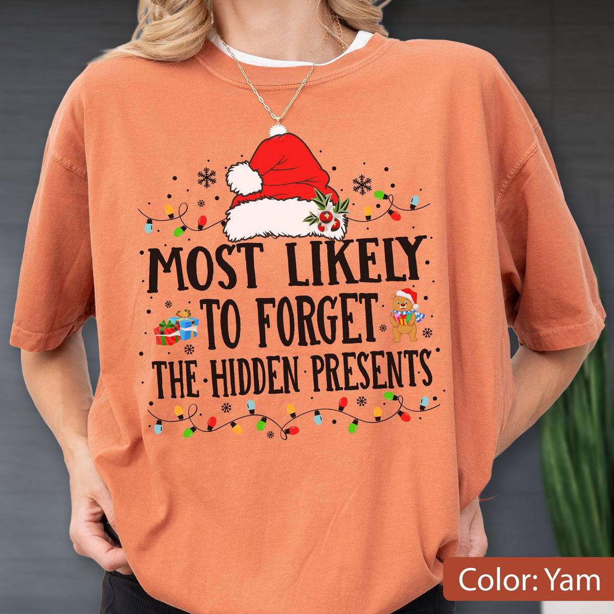 May include: A yam-colored t-shirt with a Christmas-themed graphic. The design includes a Santa hat, snowflakes, and the text "MOST LIKELY TO FORGET THE HIDDEN PRESENTS." A string of colorful lights and a teddy bear wearing a Santa hat are also featured.