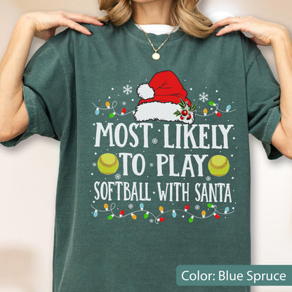 May include: A dark green t-shirt featuring a graphic with a red Santa hat, two yellow softballs, and colorful Christmas lights. The text reads: MOST LIKELY TO PLAY SOFTBALL WITH SANTA. The design includes white snowflakes and holly accents.