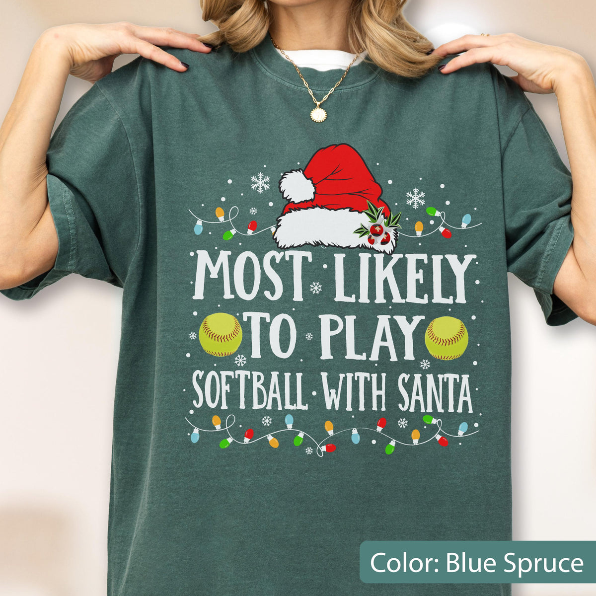 May include: Blue spruce-colored t-shirt with the text "MOST LIKELY TO PLAY SOFTBALL WITH SANTA." The design includes a Santa hat, softballs, snowflakes, and Christmas lights. A festive holiday-themed tee.