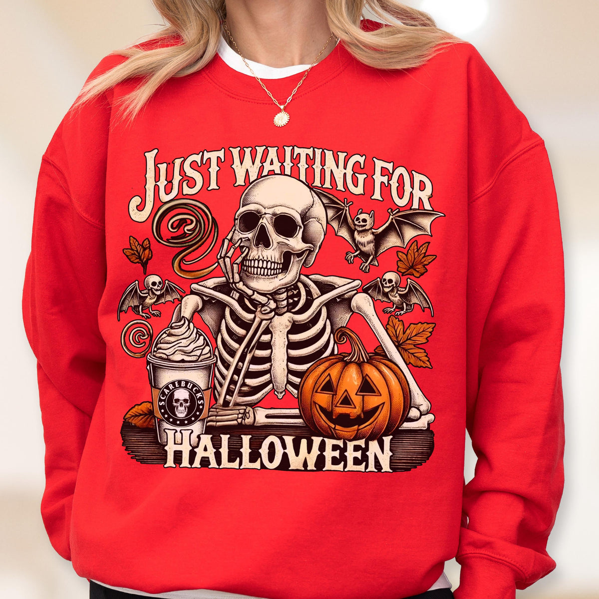 May include: A bright red crewneck sweatshirt featuring a graphic print of a skeleton resting its chin on its hand. The design includes a pumpkin, bats, autumn leaves, and a coffee cup with the text "SCAREBUCKS" and "JUST WAITING FOR HALLOWEEN".