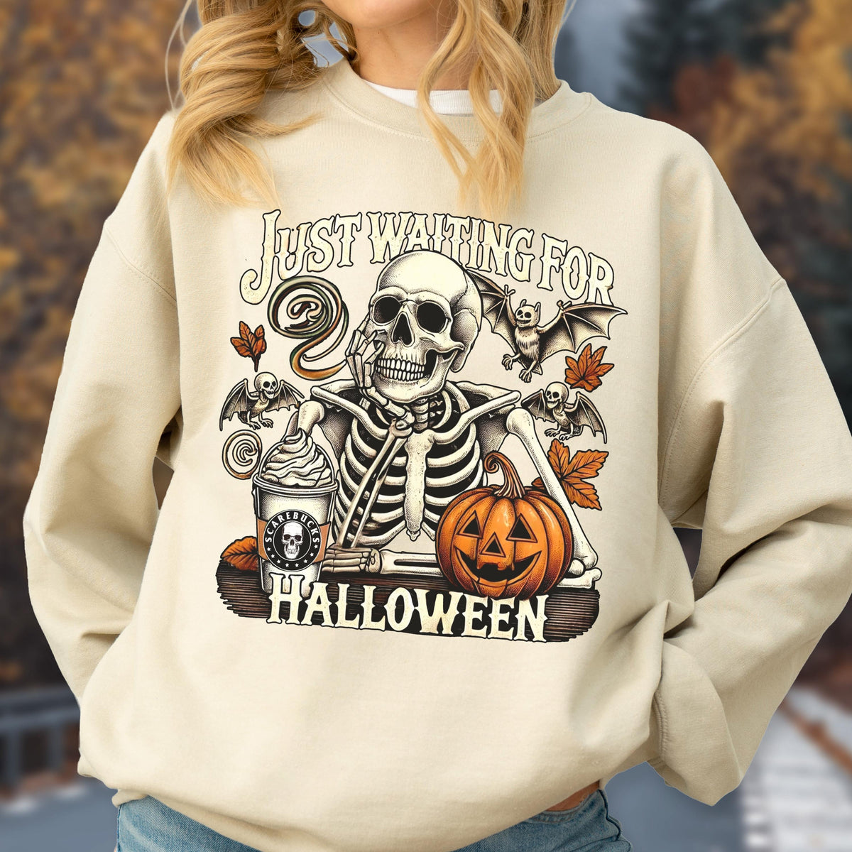 May include: A beige crewneck sweatshirt featuring a graphic print of a skeleton resting its chin on its hand. The design includes a coffee cup labeled SCARLBUCKS, a jack-o'-lantern, bats, and autumn leaves, with the text JUST WAITING FOR HALLOWEEN.