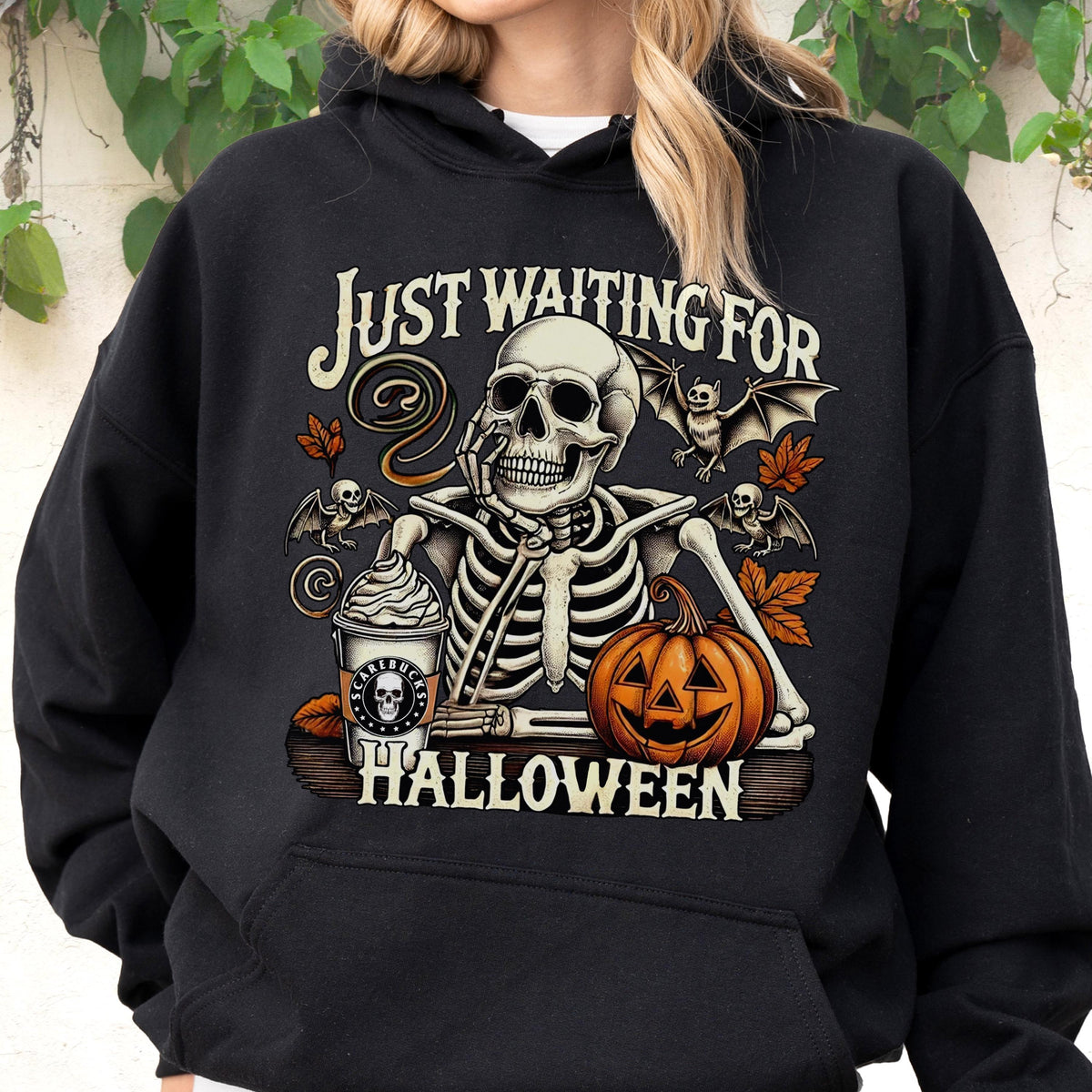 May include: A black hooded sweatshirt featuring a graphic print of a skeleton resting its chin on its hand. The design includes a cup labeled Scarebucks, a jack-o'-lantern, bats, and autumn leaves, with the text Just Waiting For Halloween.