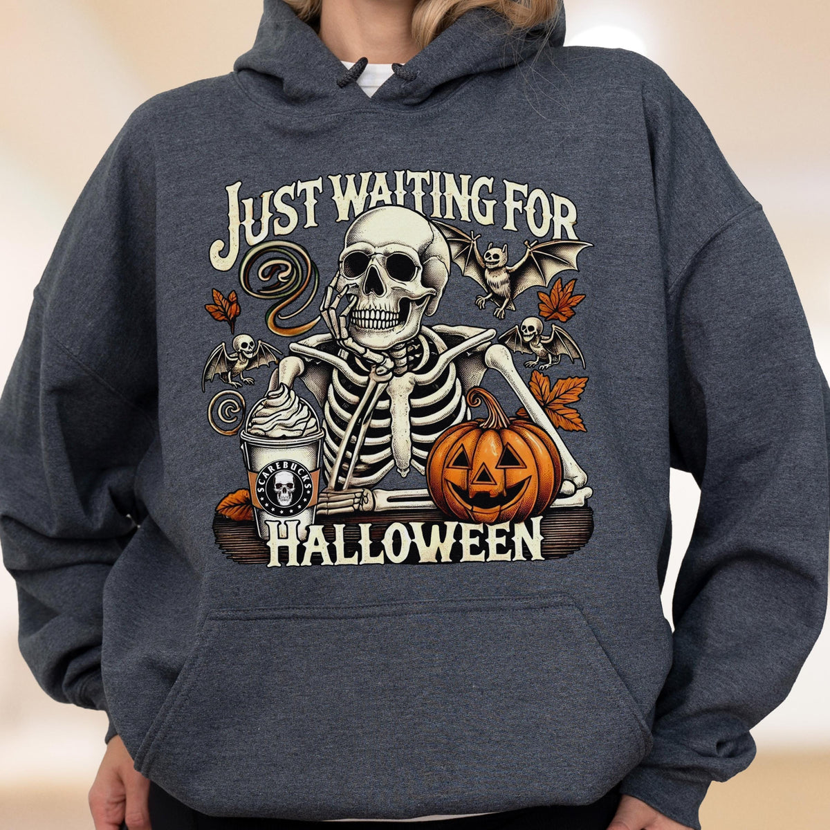 May include: A dark heather gray hooded sweatshirt featuring a graphic of a skeleton resting its chin on its hand. The design includes a coffee cup, a jack-o'-lantern, bats, and autumn leaves, with the text "JUST WAITING FOR HALLOWEEN" printed.