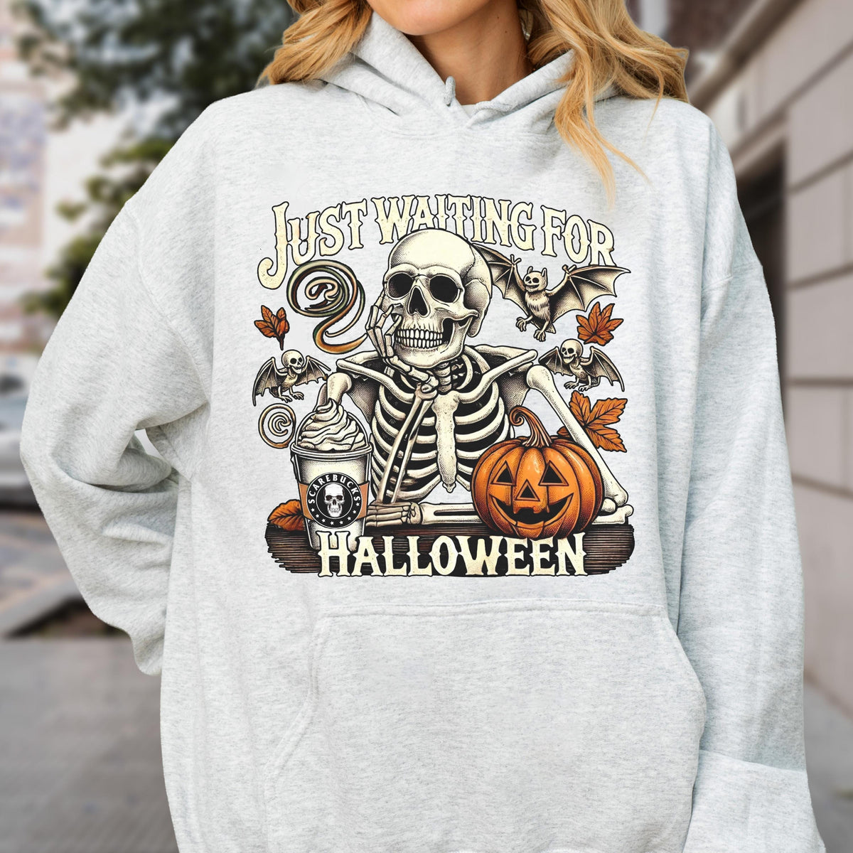 May include: A light gray hooded sweatshirt featuring a graphic print of a skeleton resting its chin on its hand next to a pumpkin and a coffee cup. The text reads "JUST WAITING FOR HALLOWEEN" with illustrations of bats and autumn leaves.