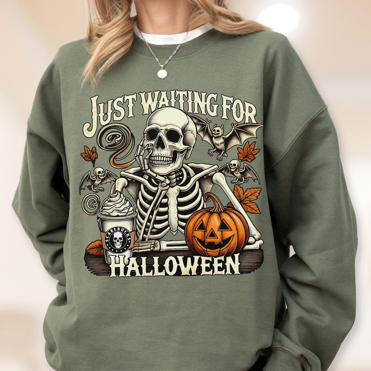 May include: An olive green crewneck sweatshirt featuring a graphic of a skeleton resting its chin on its hand. The design includes a pumpkin, bats, autumn leaves, and a coffee cup with the text "SCAREBUCKS" and "JUST WAITING FOR HALLOWEEN".