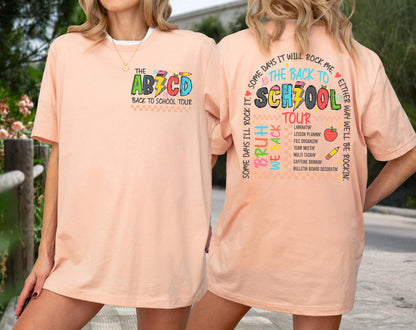 May include: Peach-colored t-shirt with the text "THE ABCD BACK TO SCHOOL TOUR" on the front. The back of the shirt features the words "THE BACK TO SCHOOL TOUR" with additional text and graphics, including an apple and pencil.
