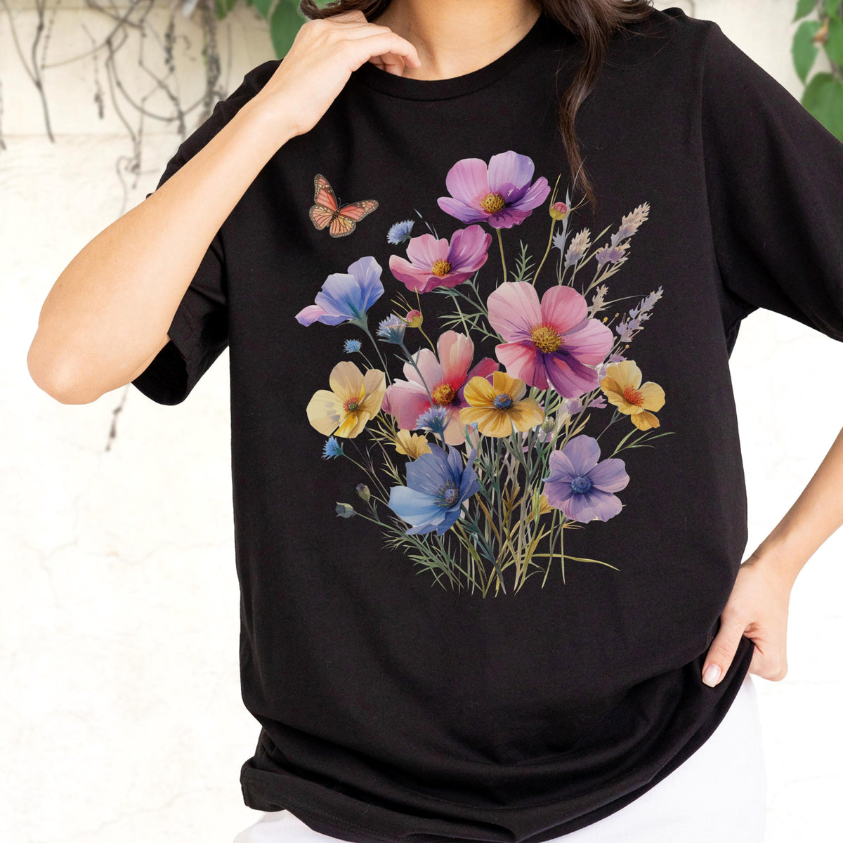 May include: A black short-sleeved t-shirt featuring a central graphic print of a colorful wildflower bouquet. The floral arrangement includes shades of pink, purple, blue, and yellow blossoms with green stems and a single orange butterfly above.