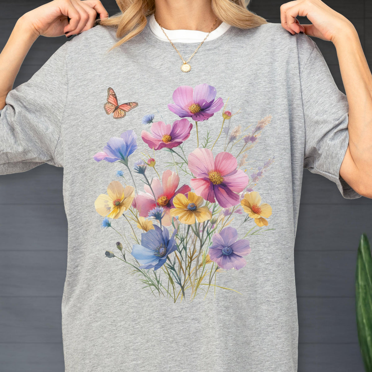 May include: A heather gray short-sleeve t-shirt featuring a colorful graphic print of a wildflower bouquet in shades of pink, yellow, blue, and purple, accompanied by a single orange butterfly positioned to the upper left of the floral arrangement.