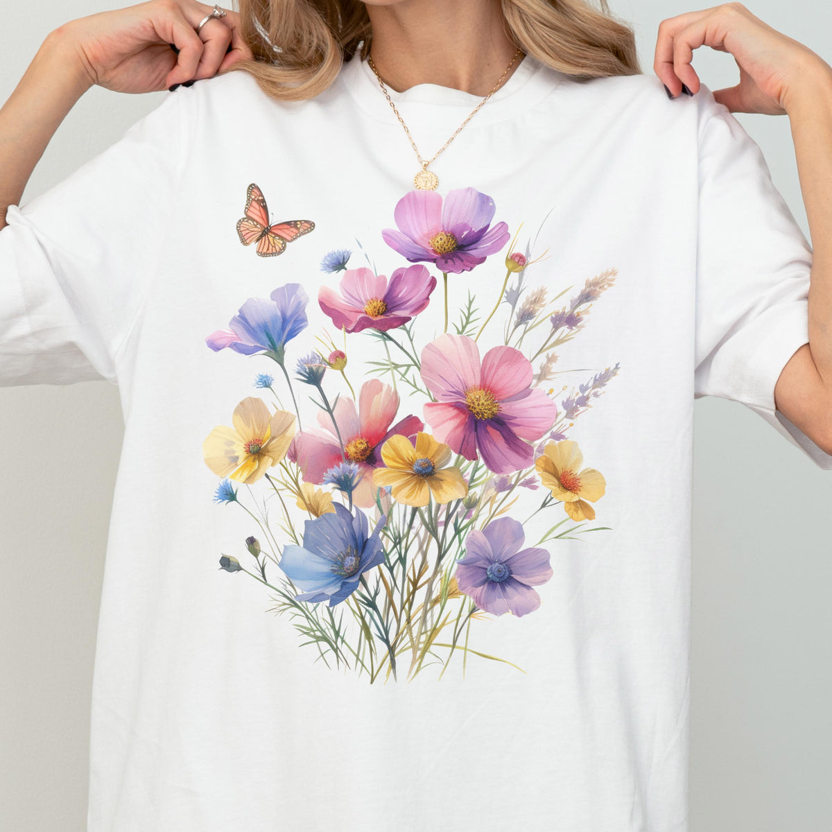 May include: A white short-sleeved t-shirt featuring a colorful graphic print of various wildflowers in shades of pink, purple, blue, and yellow, accompanied by a single orange butterfly positioned near the top left of the floral arrangement.