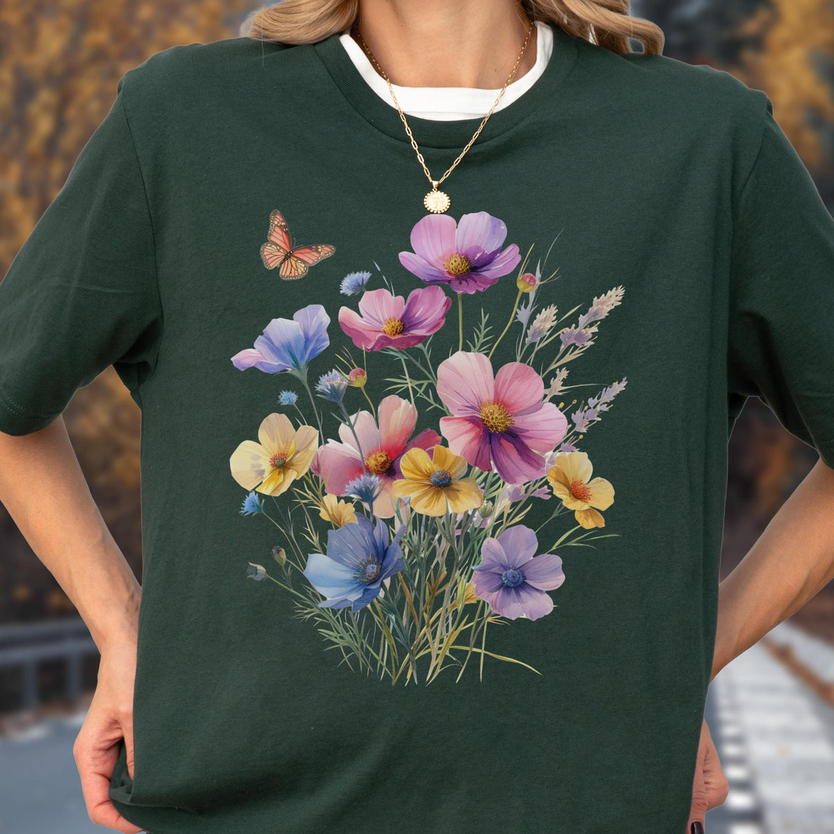 May include: A dark forest green t-shirt featuring a central graphic of a colorful wildflower bouquet in shades of pink, yellow, blue, and purple, accompanied by a single orange butterfly hovering near the top left of the floral arrangement.